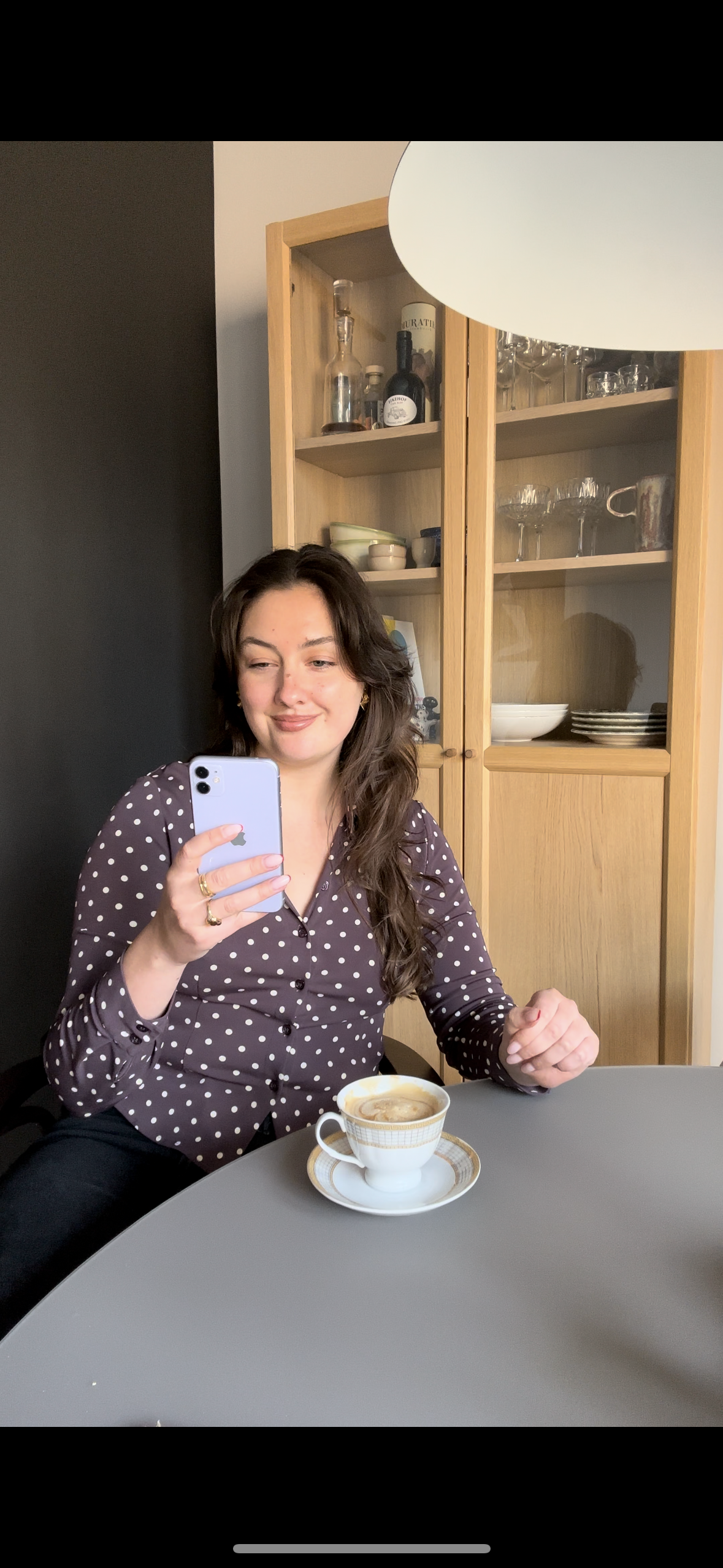 Woman sitting at a table with a cup of coffee, using a phone in a home setting.