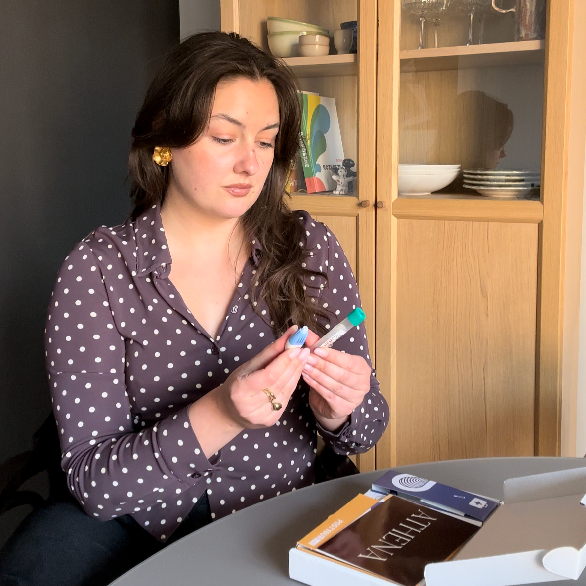 Woman sitting at a table doing the finger prick for an at-home fertility test in a room with wooden shelves.