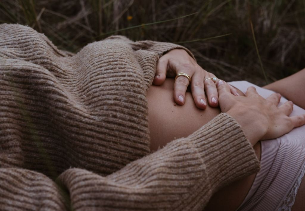 Person wearing a brown sweater holding their pregnant belly outdoors.